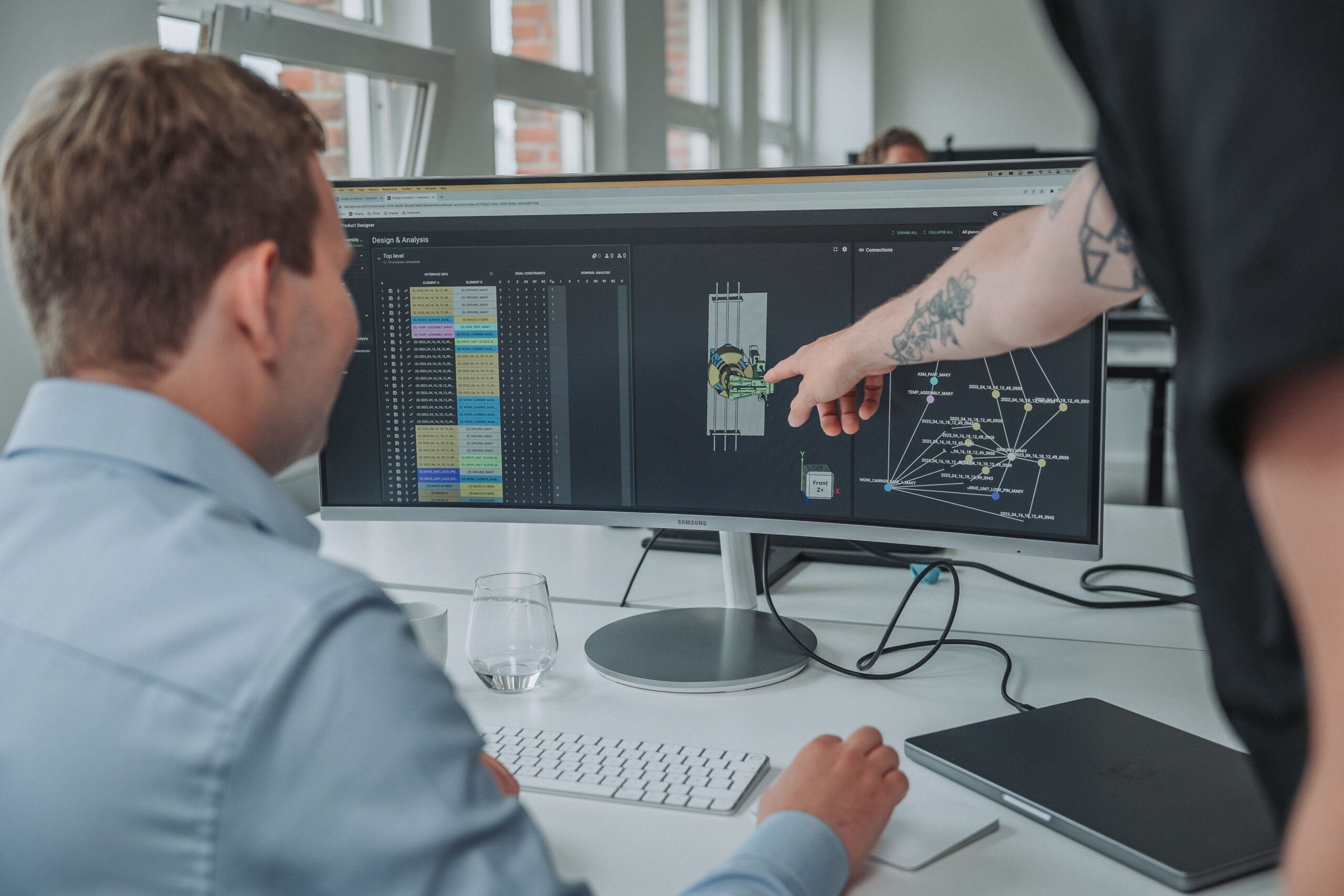 Two men working on a 3D design and data analysis displayed on a wide computer monitor in a modern office.