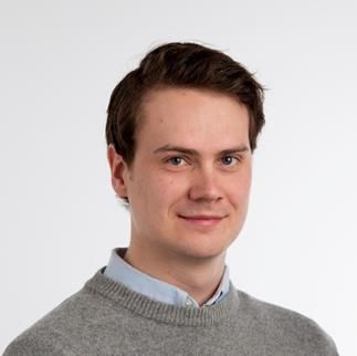 Young man with short brown hair wearing a grey sweater and blue collared shirt against a plain white background.