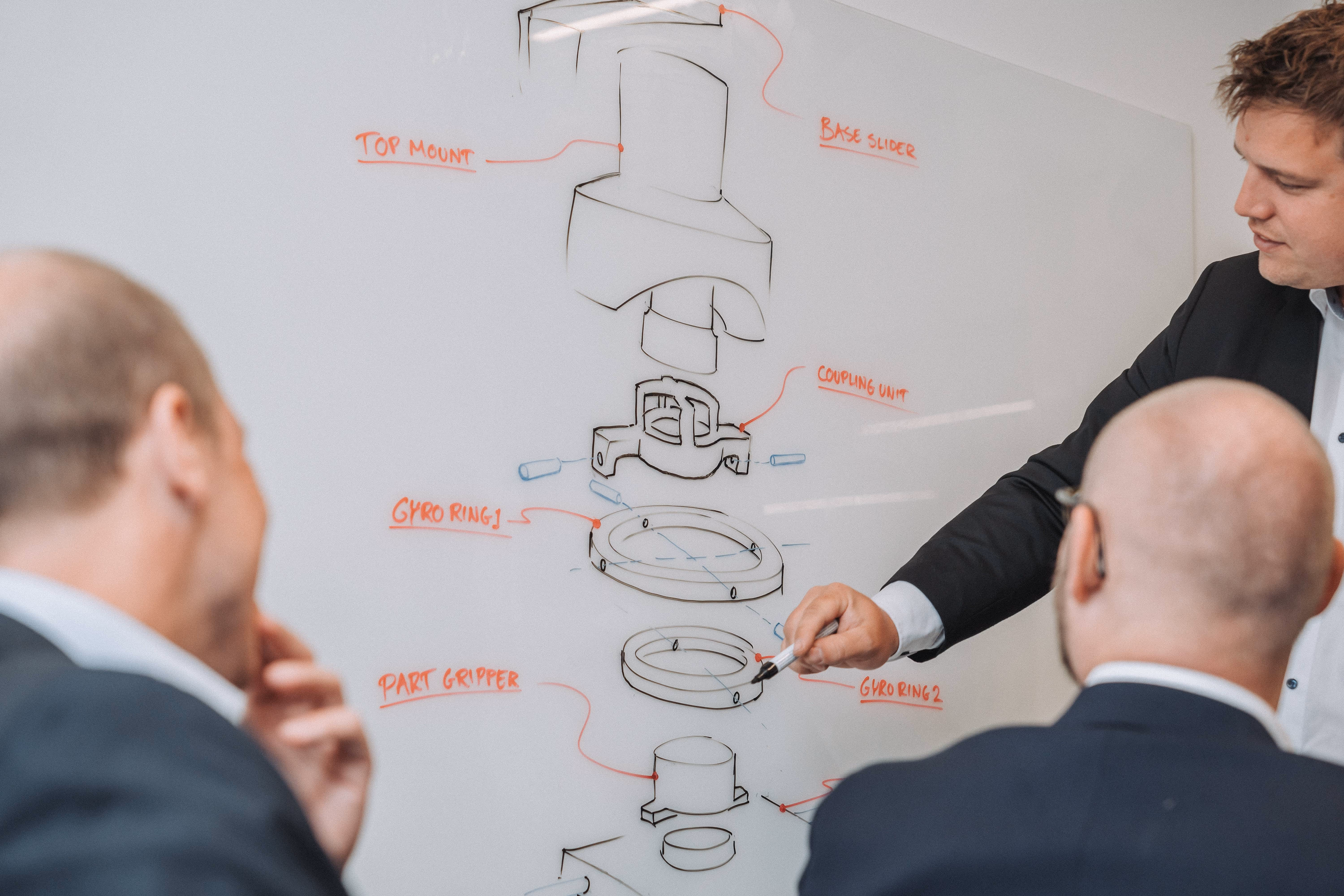 Three men in suits discussing a technical diagram labeled with component names on a whiteboard.