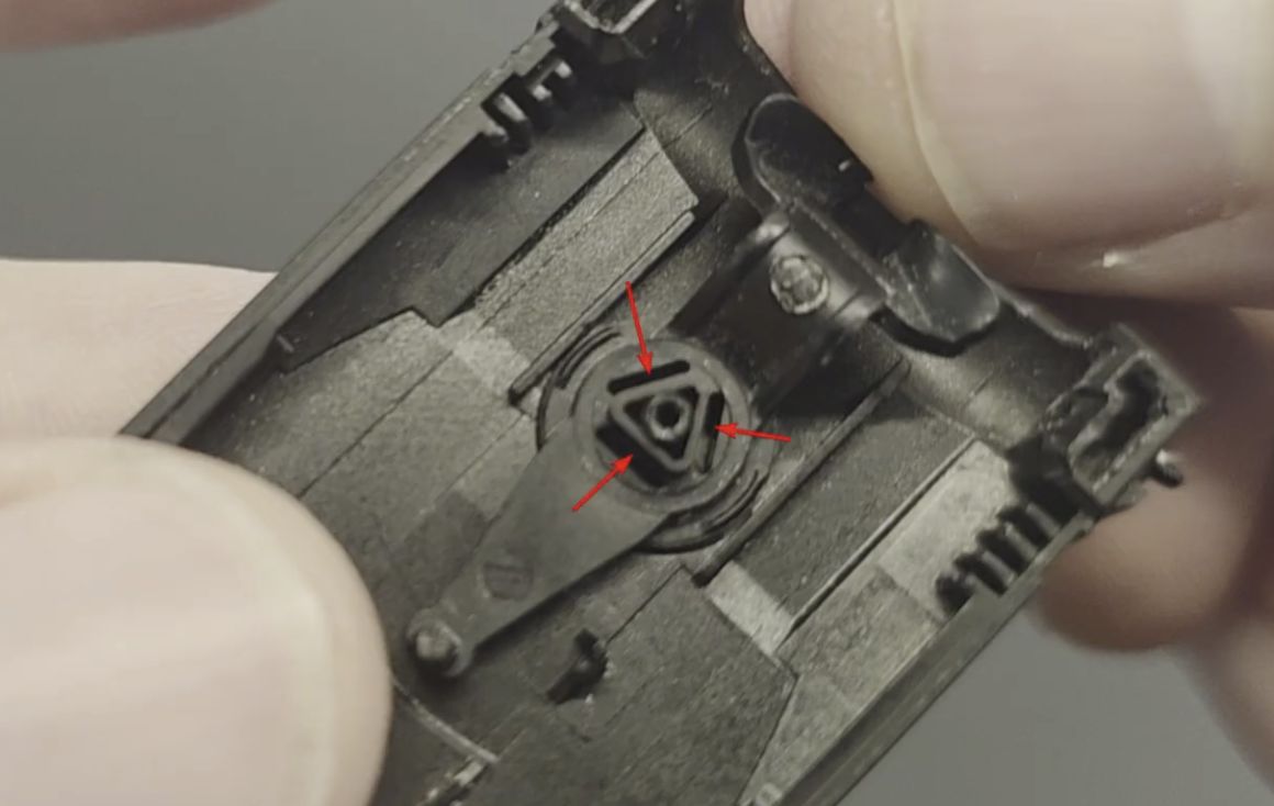 Close-up of a person's fingers holding a small black plastic device with a triangular mechanism marked by three red arrows.
