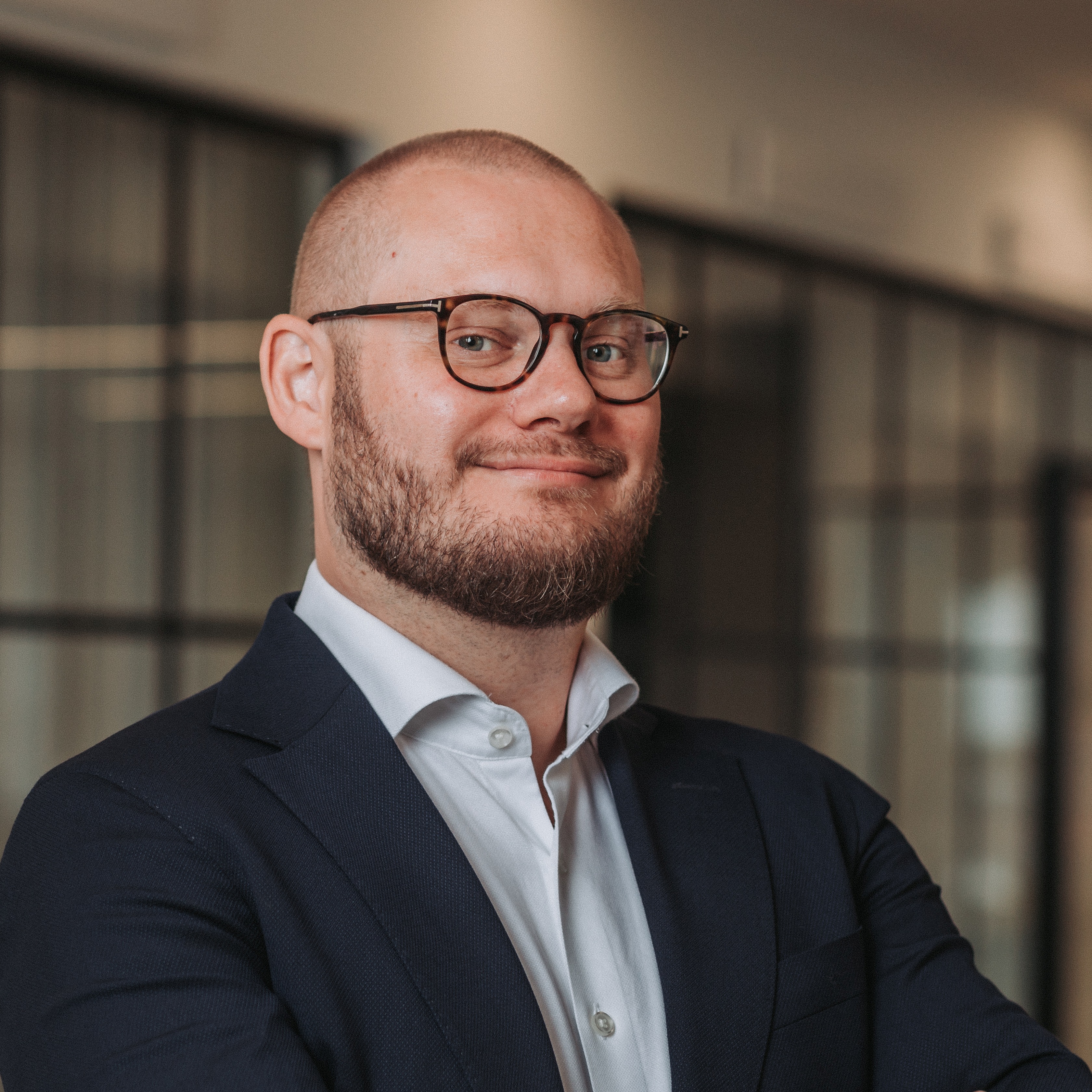 Portrait of a smiling man with a beard and glasses wearing a navy suit and white shirt in an office setting.