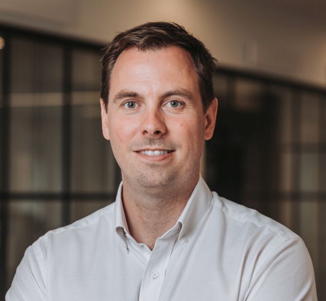 Smiling man with short brown hair wearing a white button-up shirt in an indoor office setting.