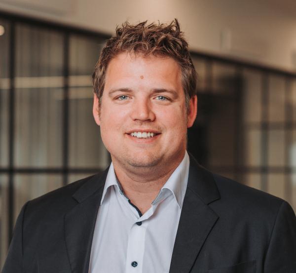 Smiling man with short brown hair wearing a dark blazer and light blue shirt in a modern office setting.
