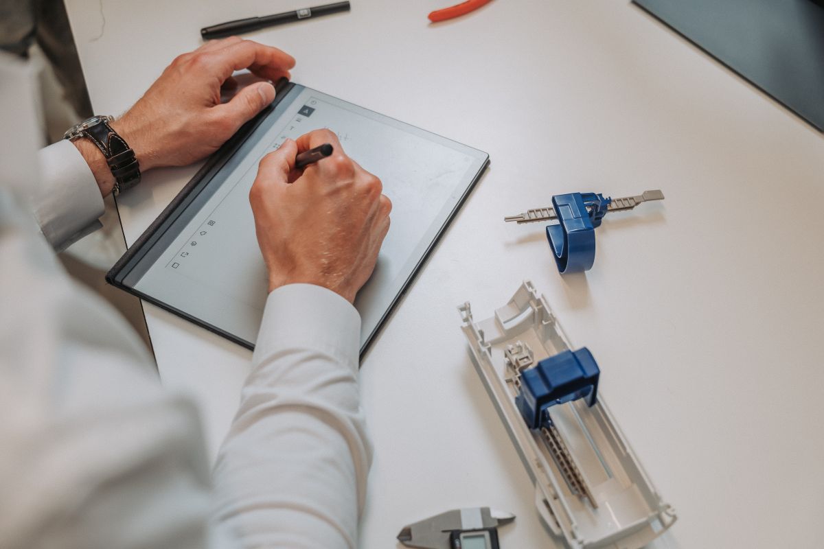 Person using a stylus to write on a tablet laying on a white desk with mechanical parts and tools nearby.