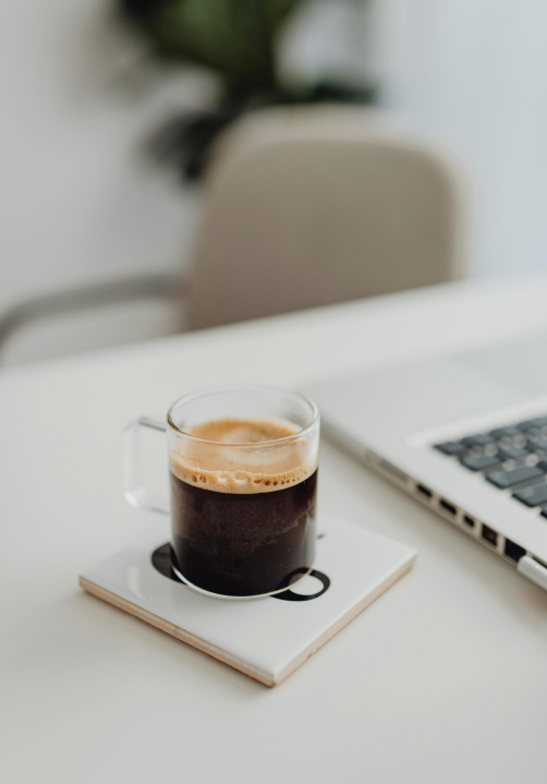 Kontakt Stimmungsbild mit einem Kaffee im Zentrum und daneben einem Laptop angeschnitten. Ruhige Atmosphäre und eher schlichte Stimmung im Bild.