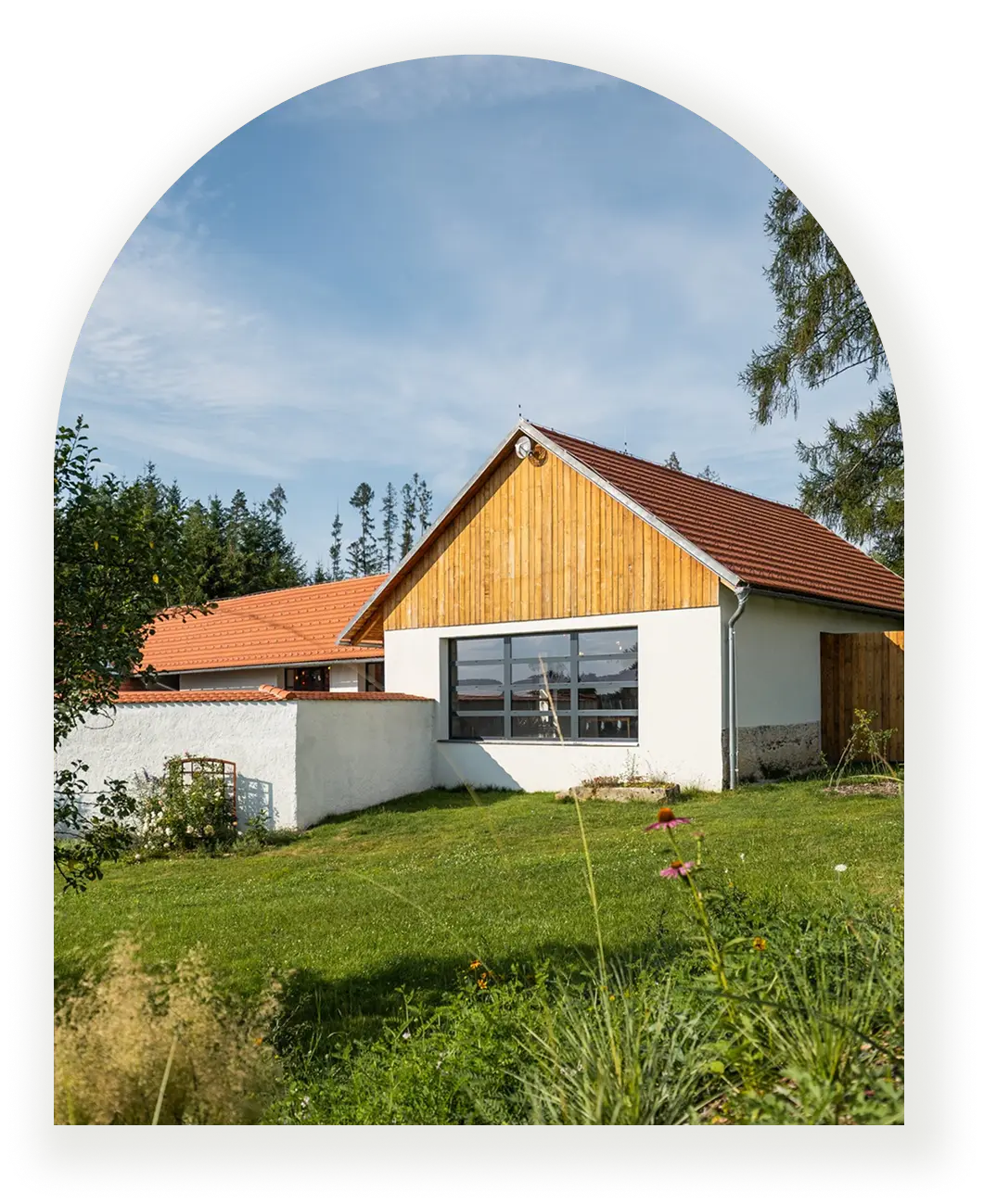 Moderní venkovský dům s bílou fasádou, dřevěným štítem a velkým oknem do zahrady.