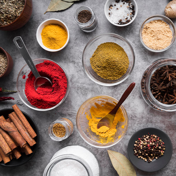 Various spices and herbs arranged in bowls and dishes on a gray surface, including cinnamon sticks, turmeric powder, star anise, mixed peppercorns, and salt.