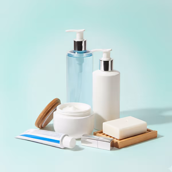 Arrangement of personal care products including two pump bottles, a jar of cream with lid, a tube, a lipstick, and a bar of soap on a wooden tray against a light blue background.