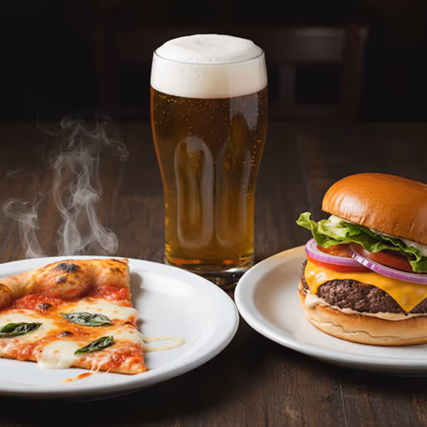 Slice of Margherita pizza with basil on a white plate, cheeseburger with lettuce, tomato, and onions on a white plate, and a pint of beer on a wooden table.