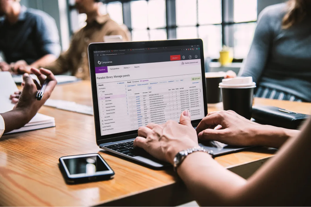 Person using a laptop displaying a panel management dashboard in an office setting with blurred people and coffee cups in the background.