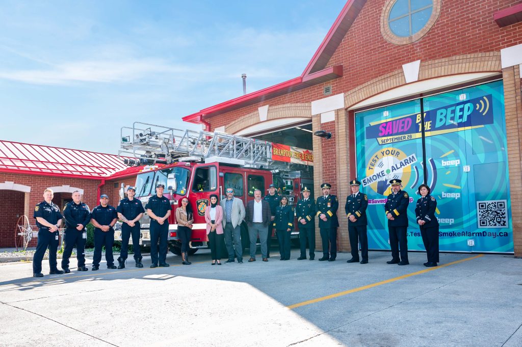 Fire service members in front of fire station bay doors. The bay doors are decorated with the "Saved by the Beep" campaign colours.
