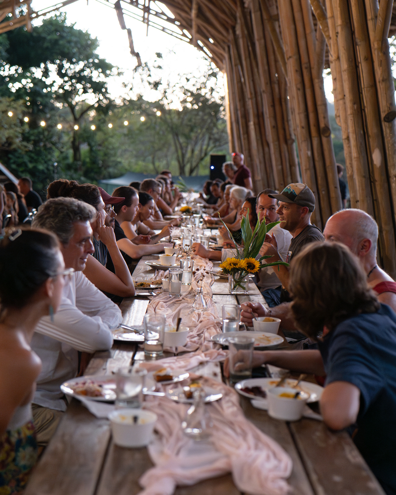 Long wooden table with people dining outdoors under a bamboo structure with string lights and a centerpiece of sunflowers.