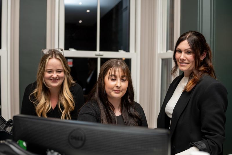 Three women smiling and working together in an office, looking at a Dell computer monitor.