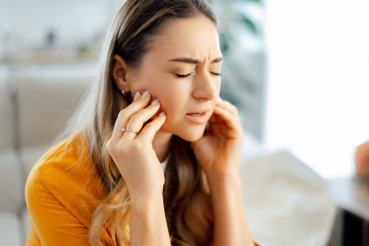 A woman sitting on a couch holding her hand to her face in discomfort.