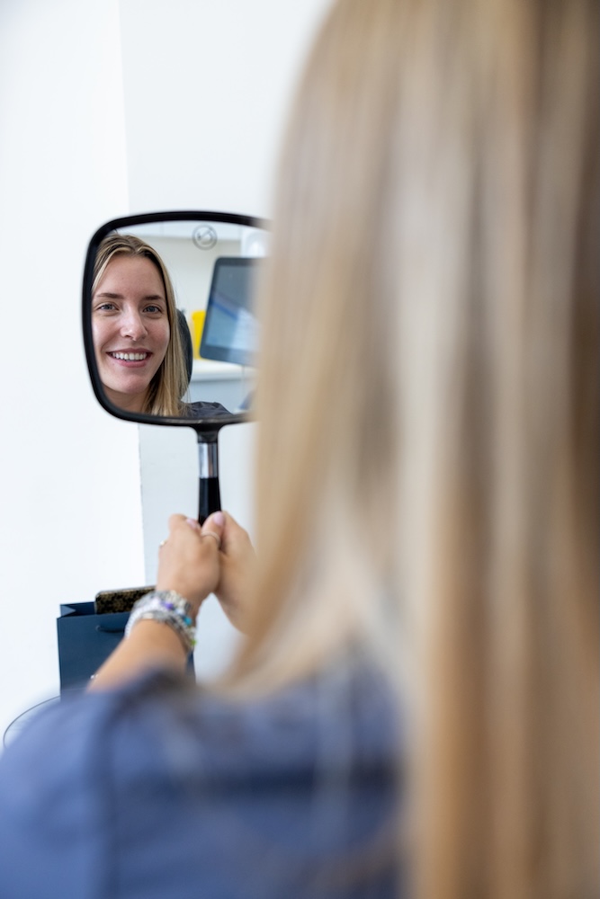 Woman holding a handheld mirror reflecting her smiling face indoors.