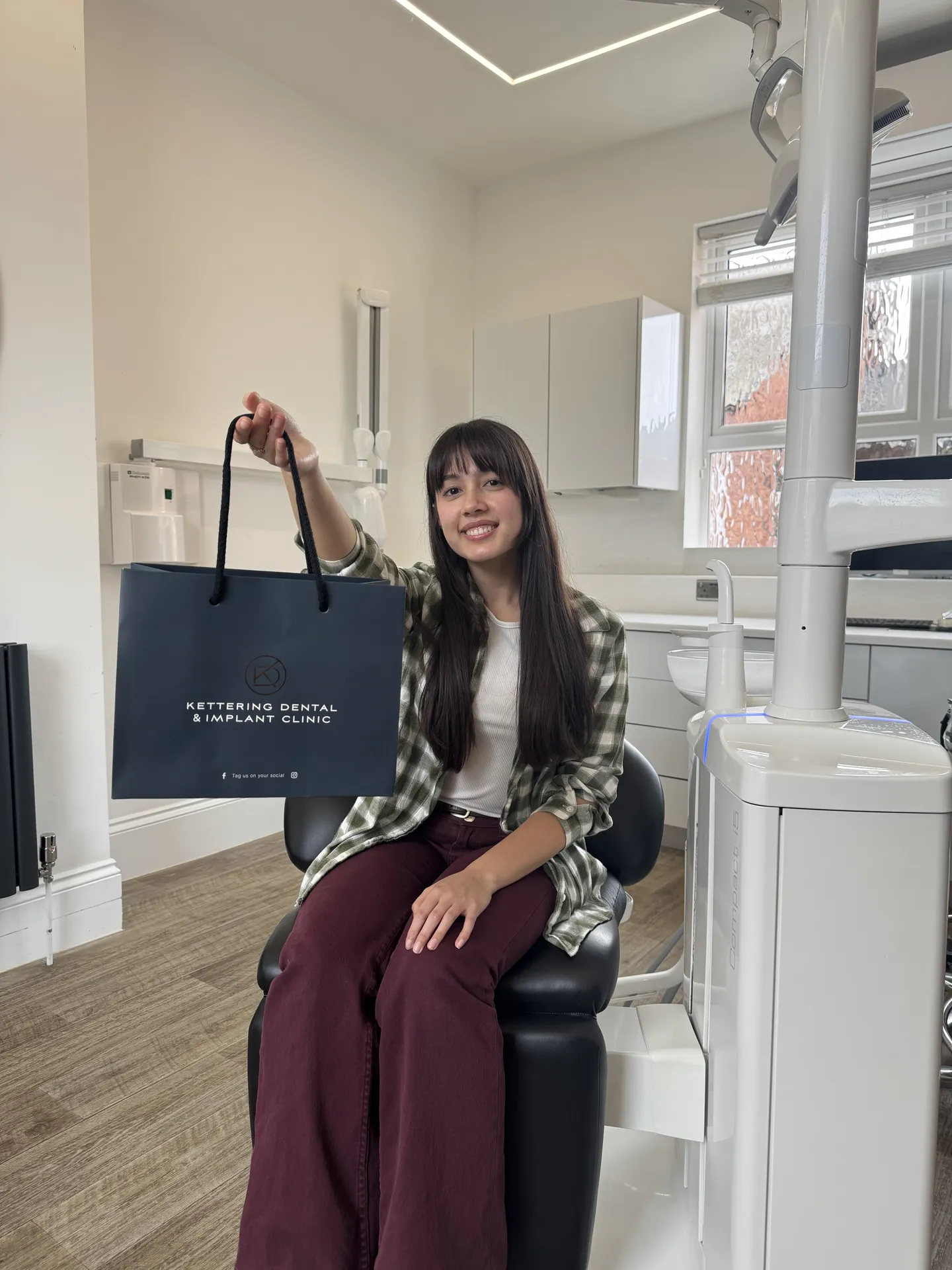 Smiling woman sitting in a dental clinic chair holding a Kettering Dental & Implant Clinic branded bag.