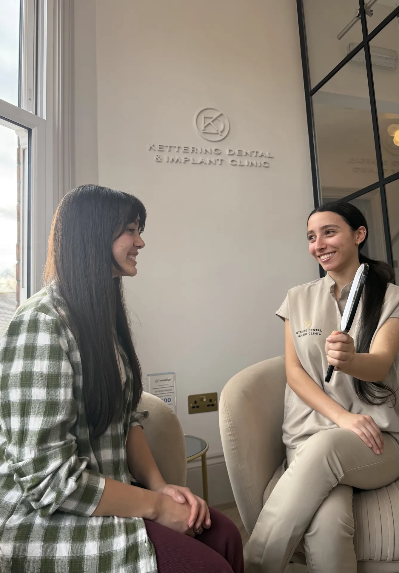 A dental professional in uniform holding a handheld mirror, smiling at a seated patient in a dental clinic waiting area.