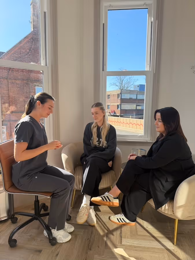 Three women seated in a sunlit room near large windows, engaged in conversation; one woman wears scrubs, the others wear casual clothes and sneakers.