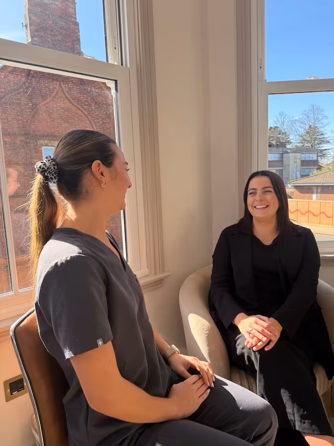 Two women sitting and smiling while having a conversation by a window with a brick building visible outside.