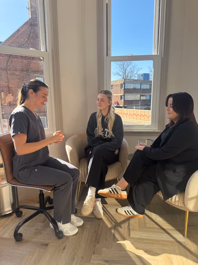 Three women sitting and talking in a sunlit room with large windows and wooden flooring.