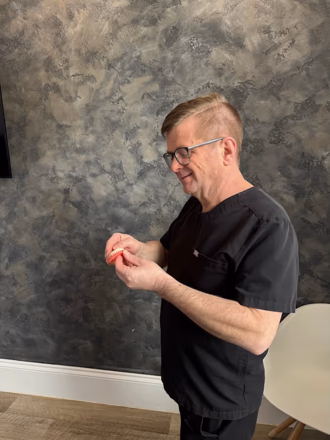 A male dentist in black scrubs holding and examining a set of dentures against a textured gray wall background.