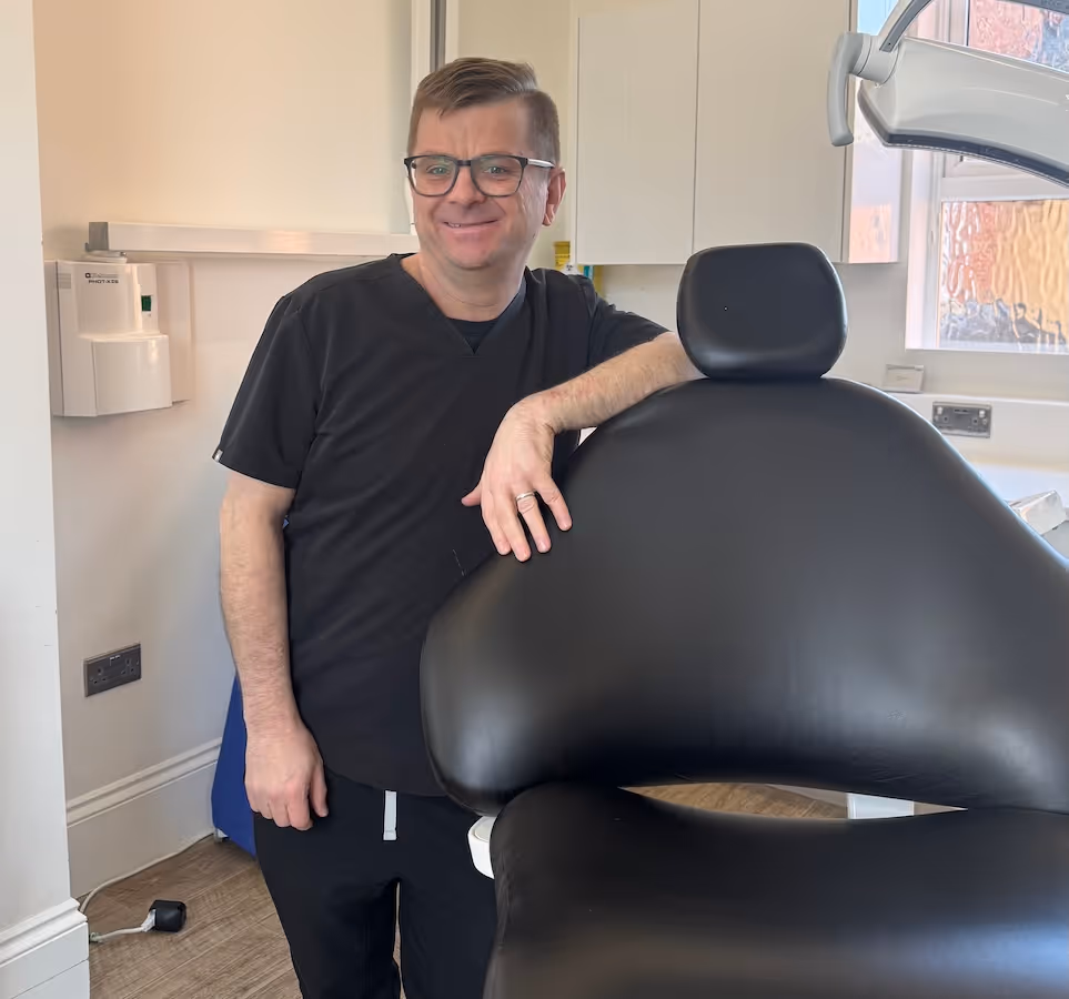 Smiling man in black scrubs leaning on a large black dental chair in a dental office.