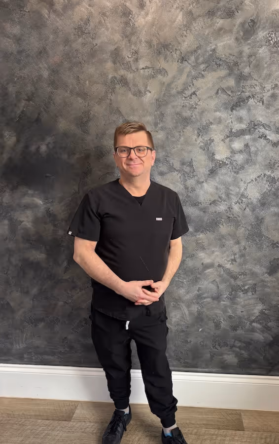 Man wearing black scrubs and glasses standing in front of a textured gray wall.