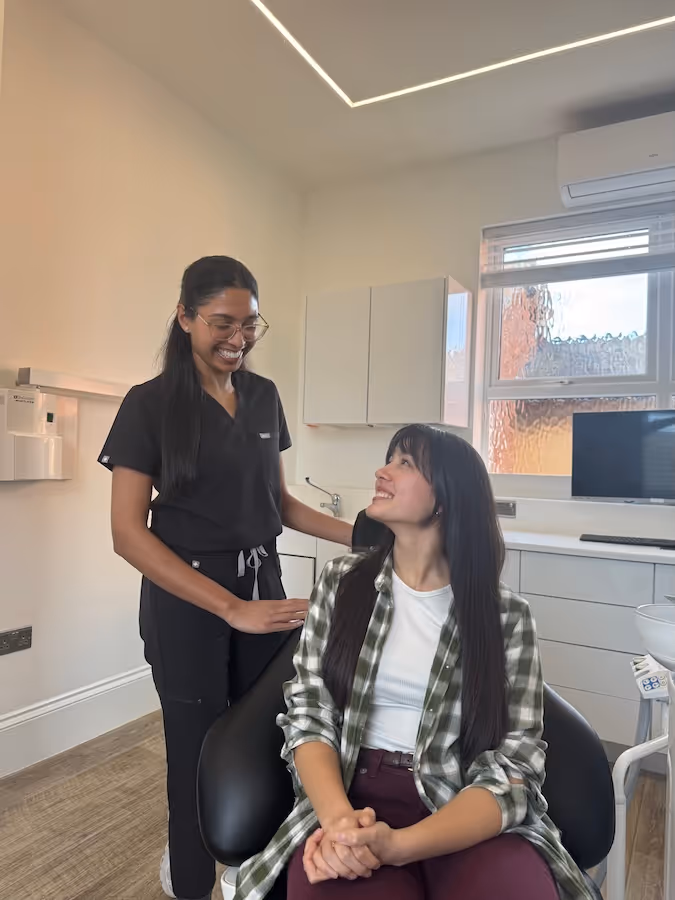 Smiling medical professional in black scrubs standing beside a seated woman in a plaid shirt in a bright examination room.