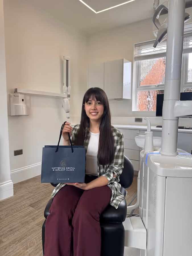 Smiling woman sitting in a dental clinic chair holding a Kettering Dental & Implant Clinic gift bag.