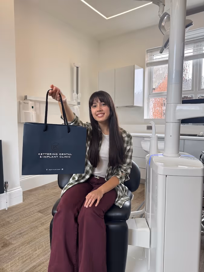 Young woman sitting on a dental chair holding a Kettering Dental & Implant Clinic branded bag inside a dental office.
