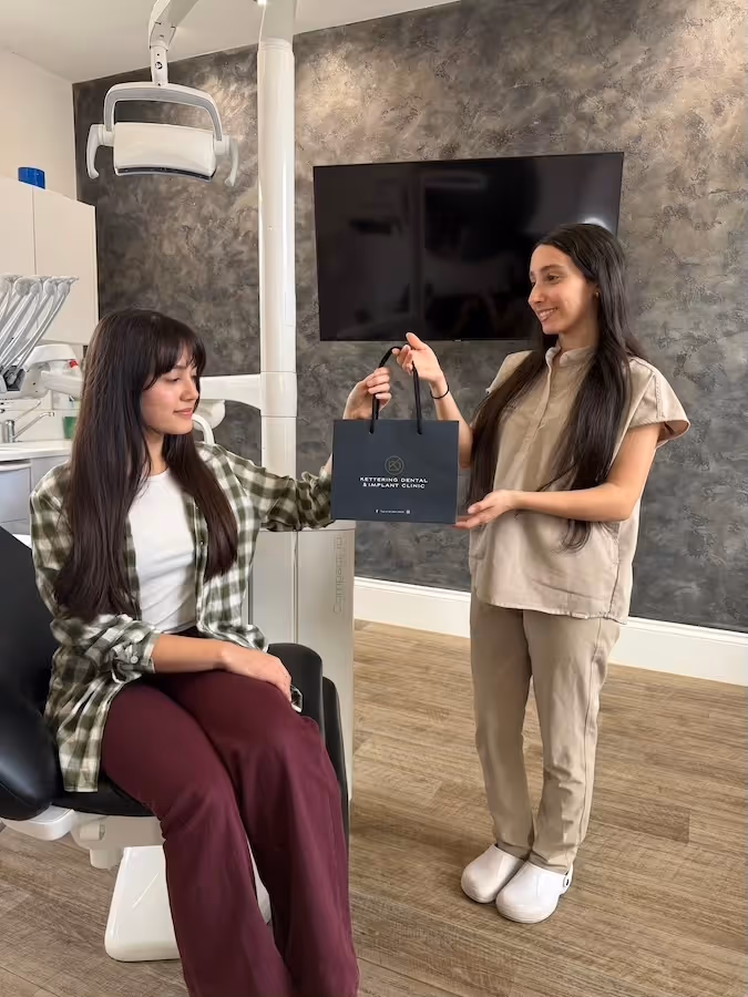 A woman sitting in a dental chair receives a black gift bag from a smiling dental clinic staff member in beige scrubs.