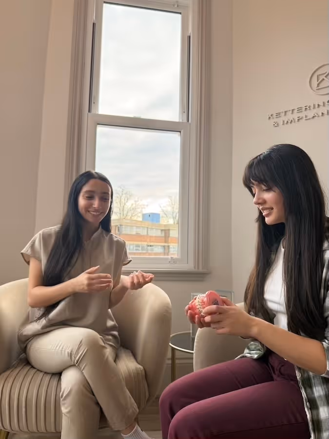 Two women seated indoors by a window, one holding a dental model, likely discussing dental care.