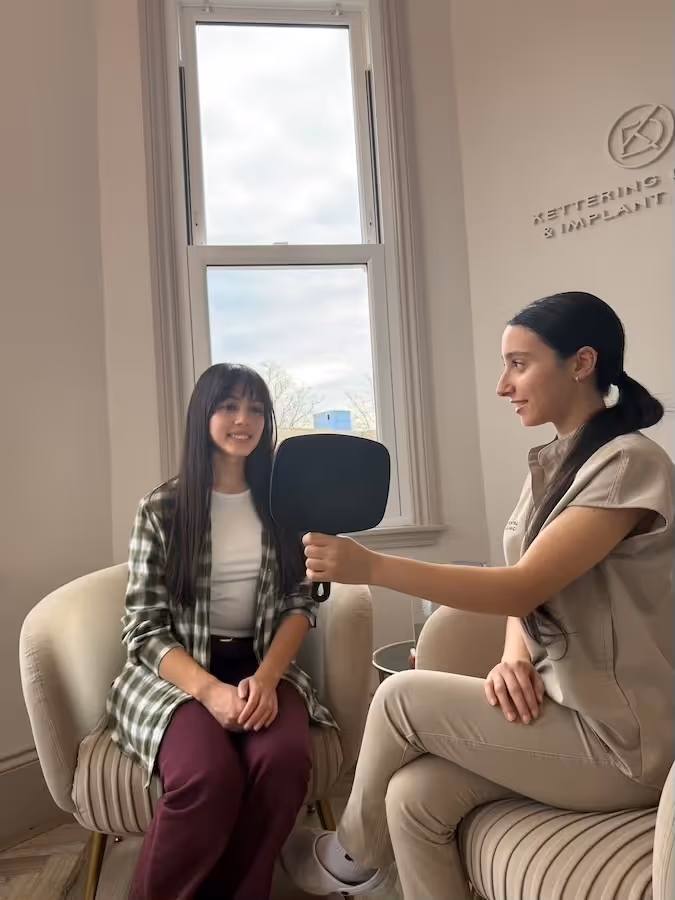 A dental professional holding a mirror as a young woman smiles sitting in a chair, with a window in the background.