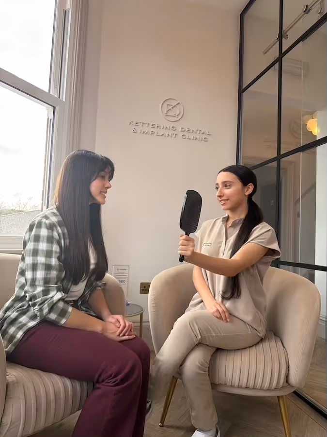 A dental professional holding a mirror and smiling while talking to a female patient seated across in a clinic waiting area.