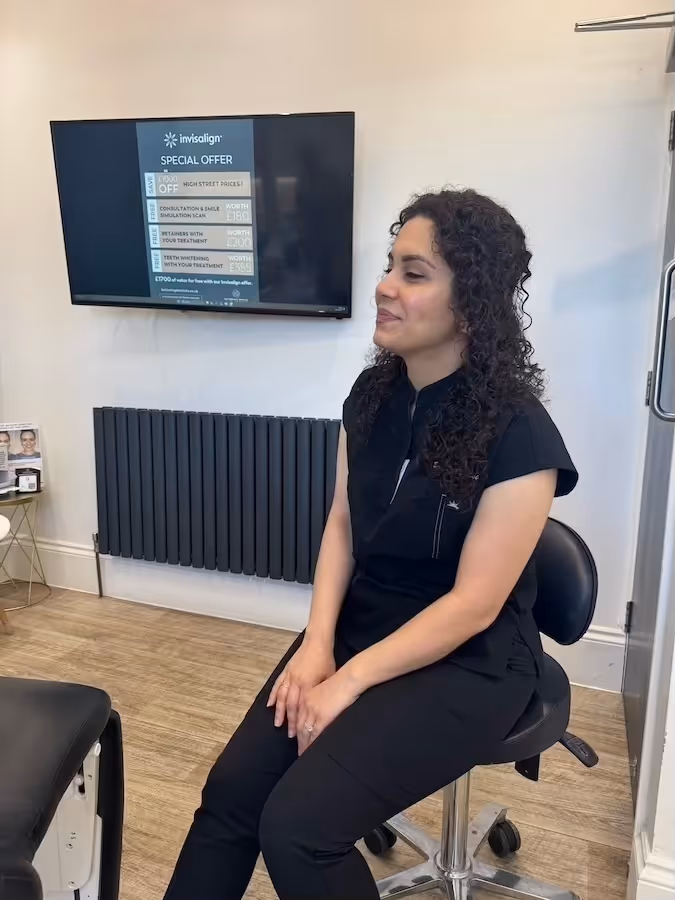 Woman with curly hair sitting on a black stool in a dental or orthodontic clinic with an Invisalign special offer display on a wall-mounted screen behind her.