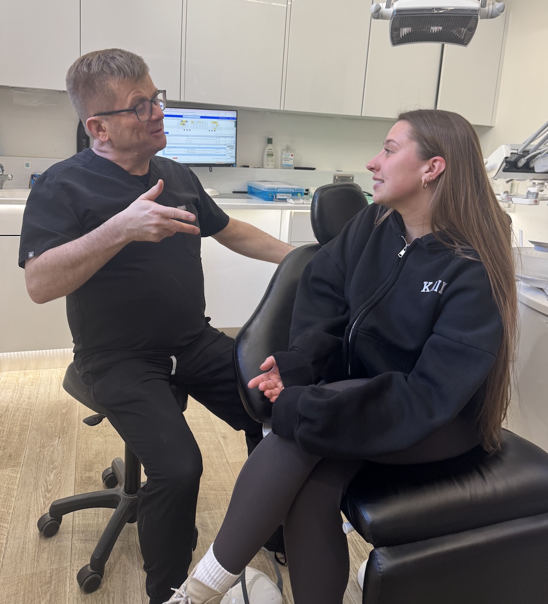 Dentist in black scrubs talking to a female patient seated in a dental chair in a clinic.