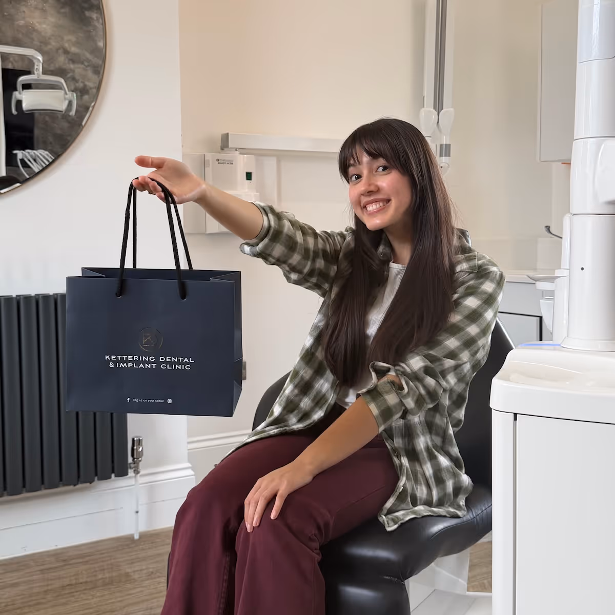 Smiling woman sitting in dental clinic chair holding a Kettering Dental & Implant Clinic bag.