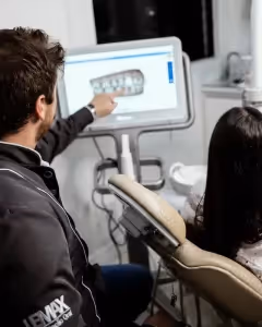 Dentist wearing dark grey uniform, pointing at the screen showing Dentures, and patient sitting on a light brown chair 