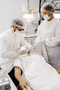 2 Dentists  wearing their professional white uniforms and hair cap, cleaning a patient's teeth. 