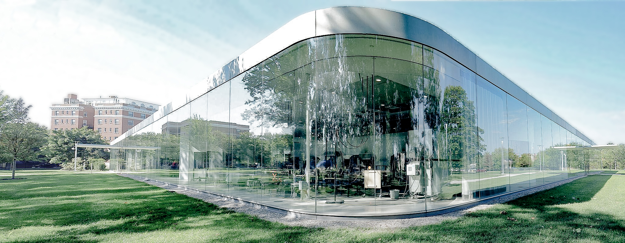 Glass-walled modern building with curved edges reflecting trees and sky, set in a green park with city buildings in the background.