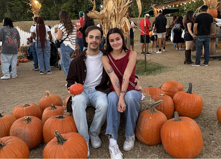 Two people sitting on a hale bale surrounded by pumpkins.