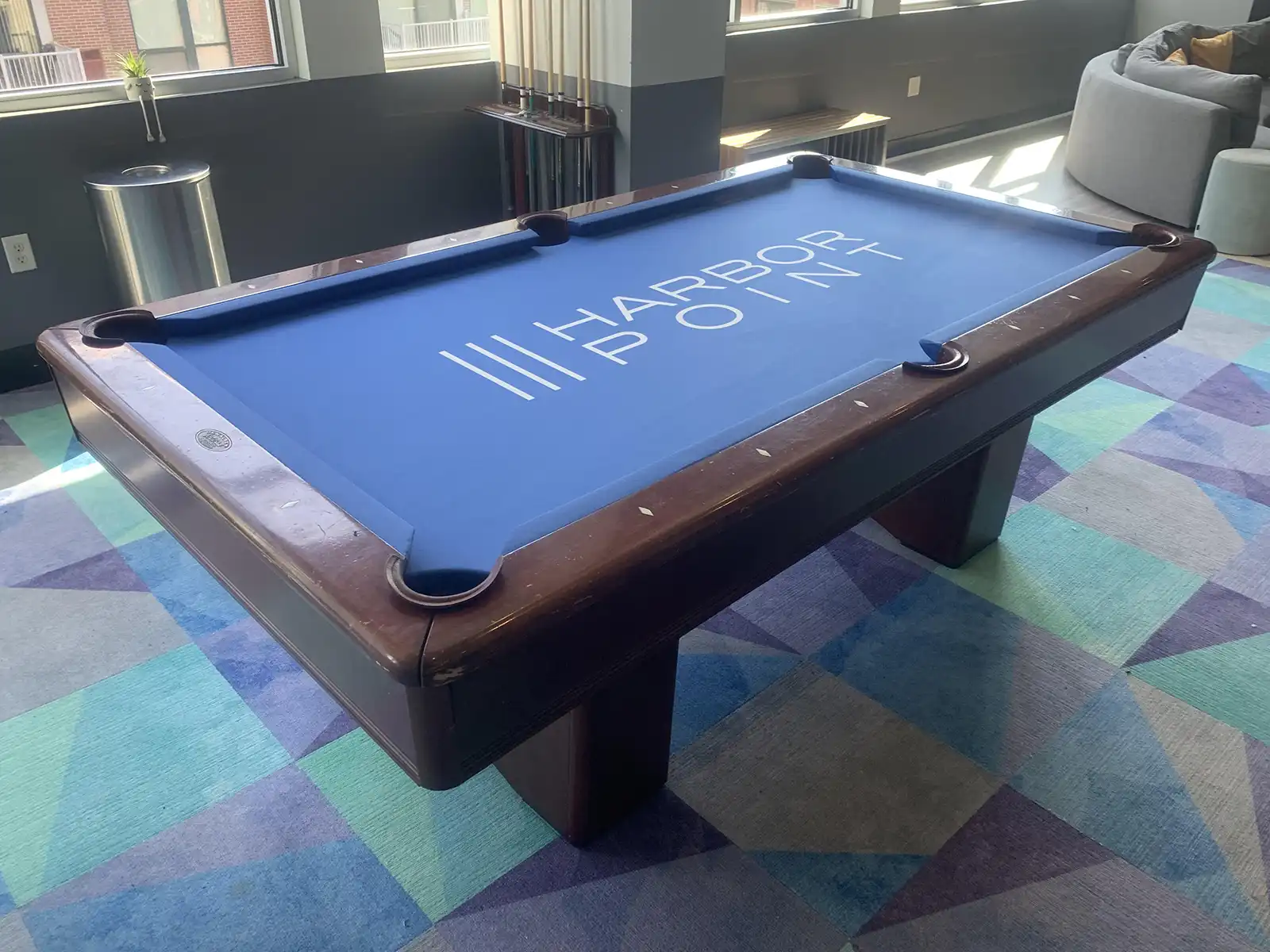 A brown pool table with a blue felt top and the logo "Harbor Point" in white. The table is in a modern, well-lit room with large windows and colorful carpeting.