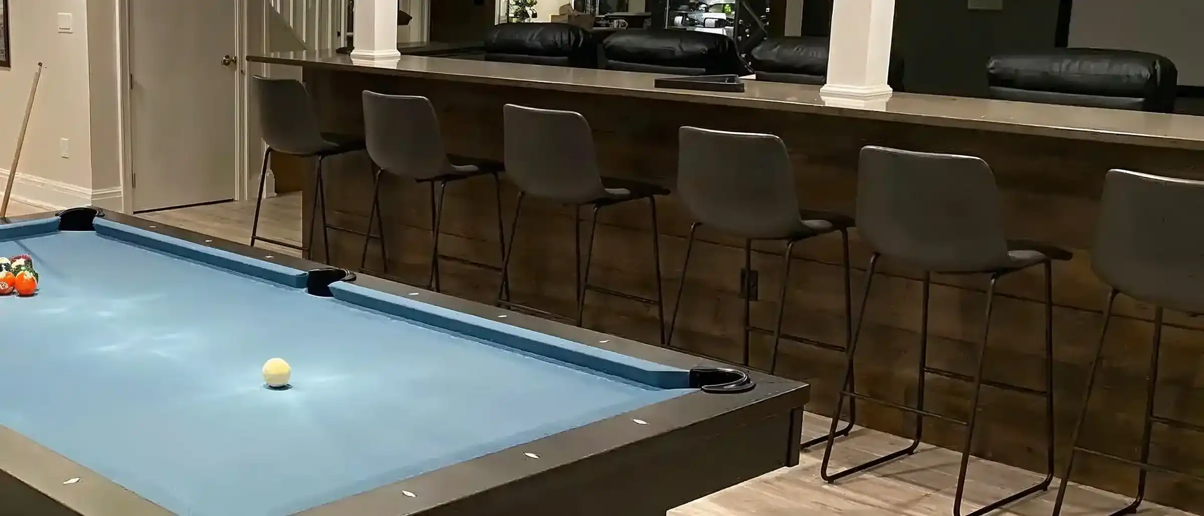 Modern basement recreation room with a light blue pool table, six gray bar stools at a wooden bar, and comfortable black leather seating in the background.