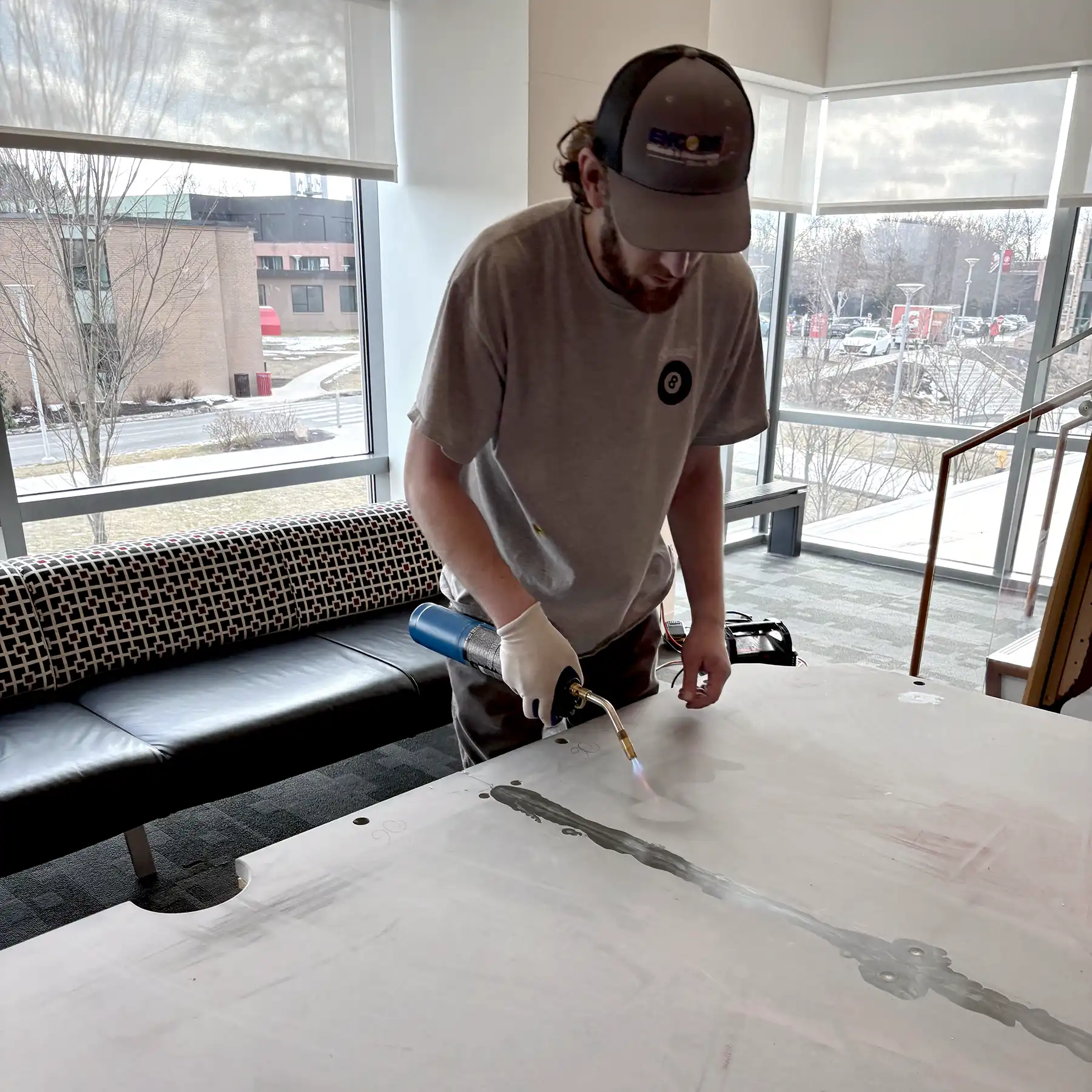 A person in a grey t-shirt and gloves uses a blowtorch to work on a large surface in a well-lit room with expansive windows. The setting is industrial and bright.