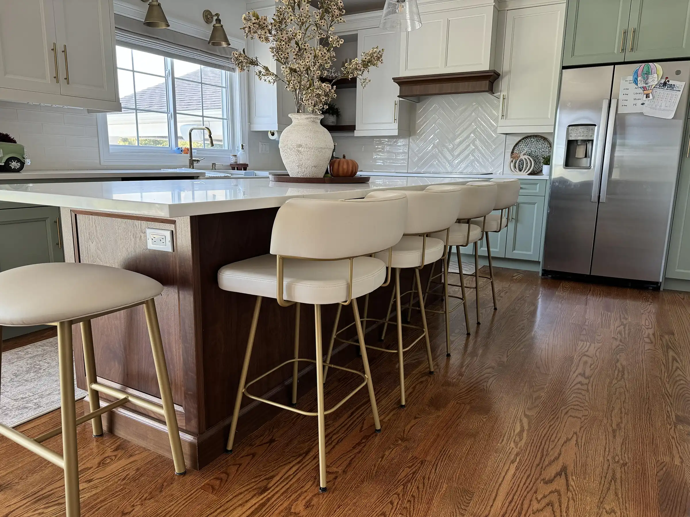 Modern kitchen with white cabinets, green accents, and a large island with upholstered counter stools. Hardwood flooring and a farmhouse sink complete the space.
