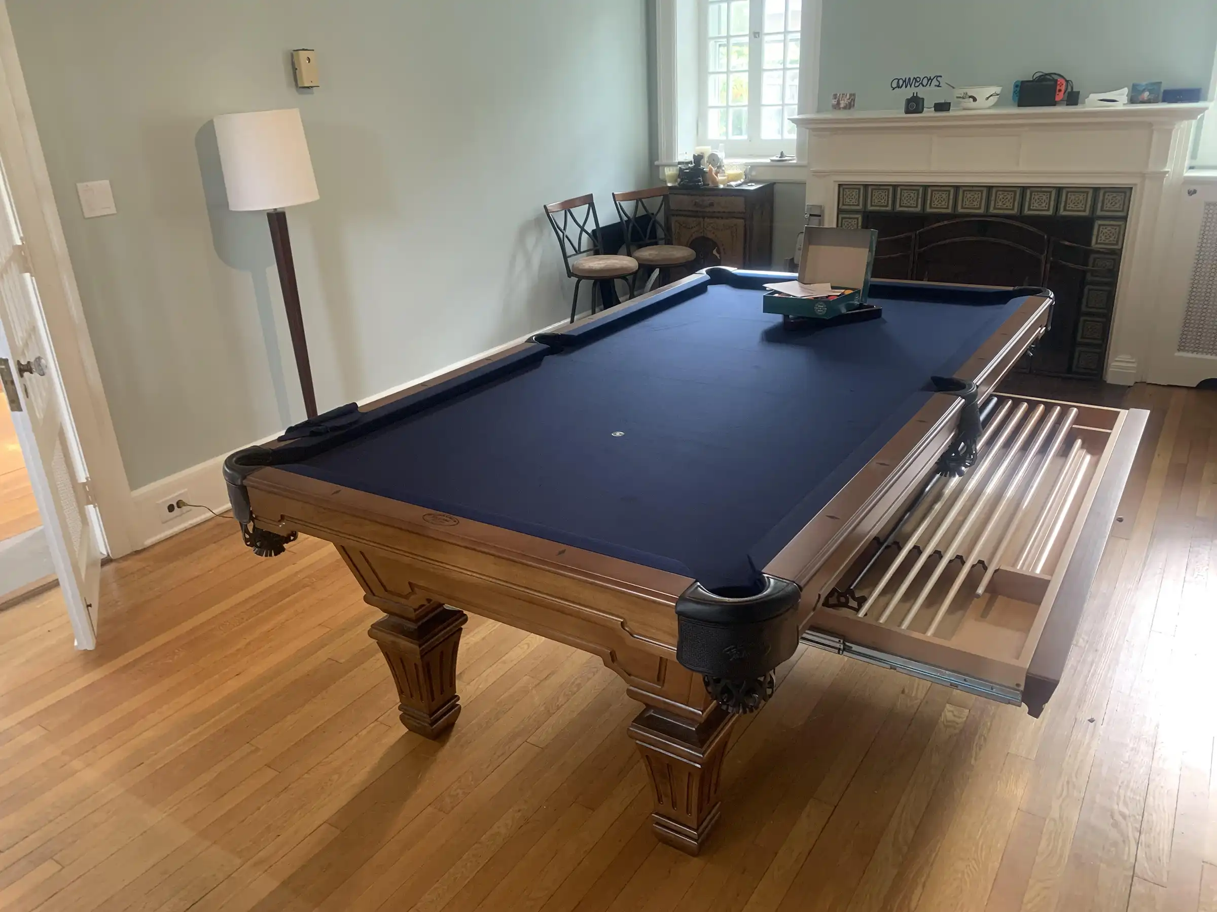An Olhausen pool table with a blue felt surface is positioned in a spacious room with hardwood floors. A fireplace with a tiled mantlepiece is visible in the background.