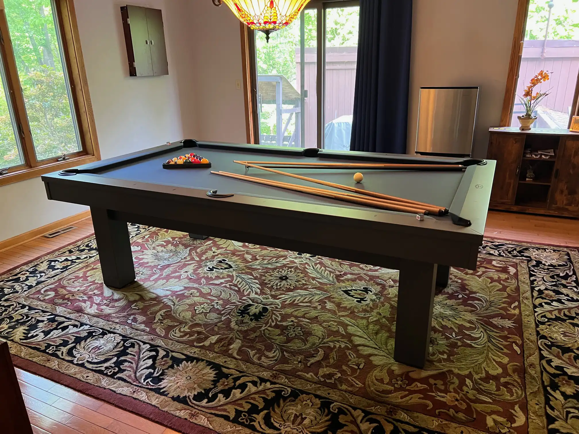 Sleek Olhausen West End pool table with a dark gray finish and blue felt, complemented by matching cues and a set of billiard balls. The table sits on a vibrant patterned rug in a bright, modern room.