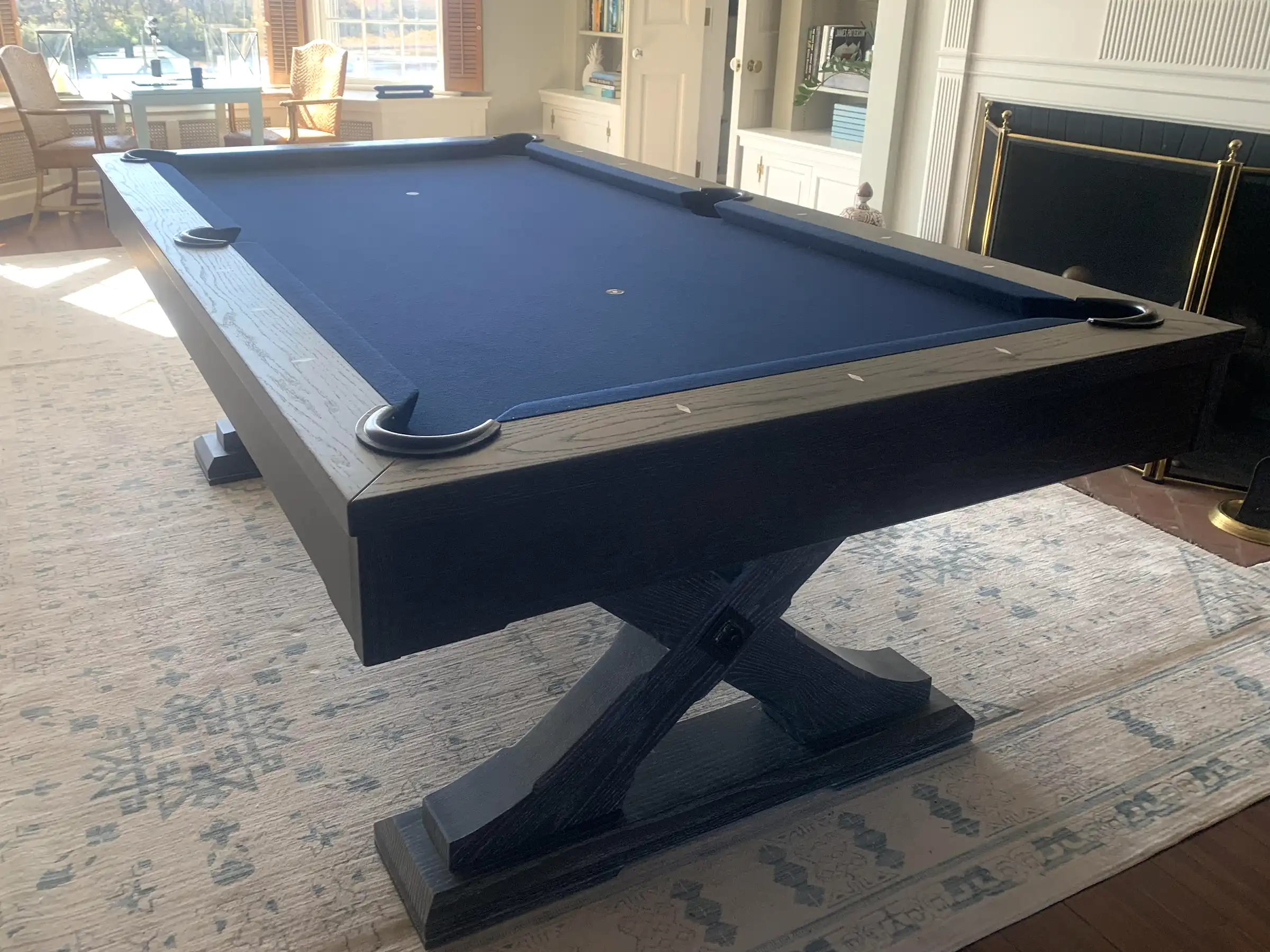 Blue felt pool table with a dark wood X-shaped base, located in a bright living room.