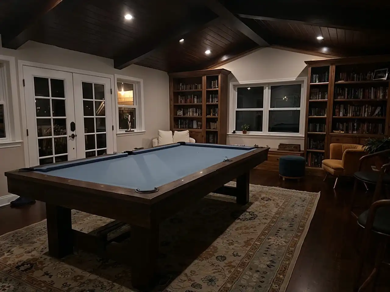 Cozy game room with blue grey felt pool table on patterned rug, featuring built-in wooden bookshelves, vaulted dark wood ceiling, French doors, and warm accent lighting for ambiance.