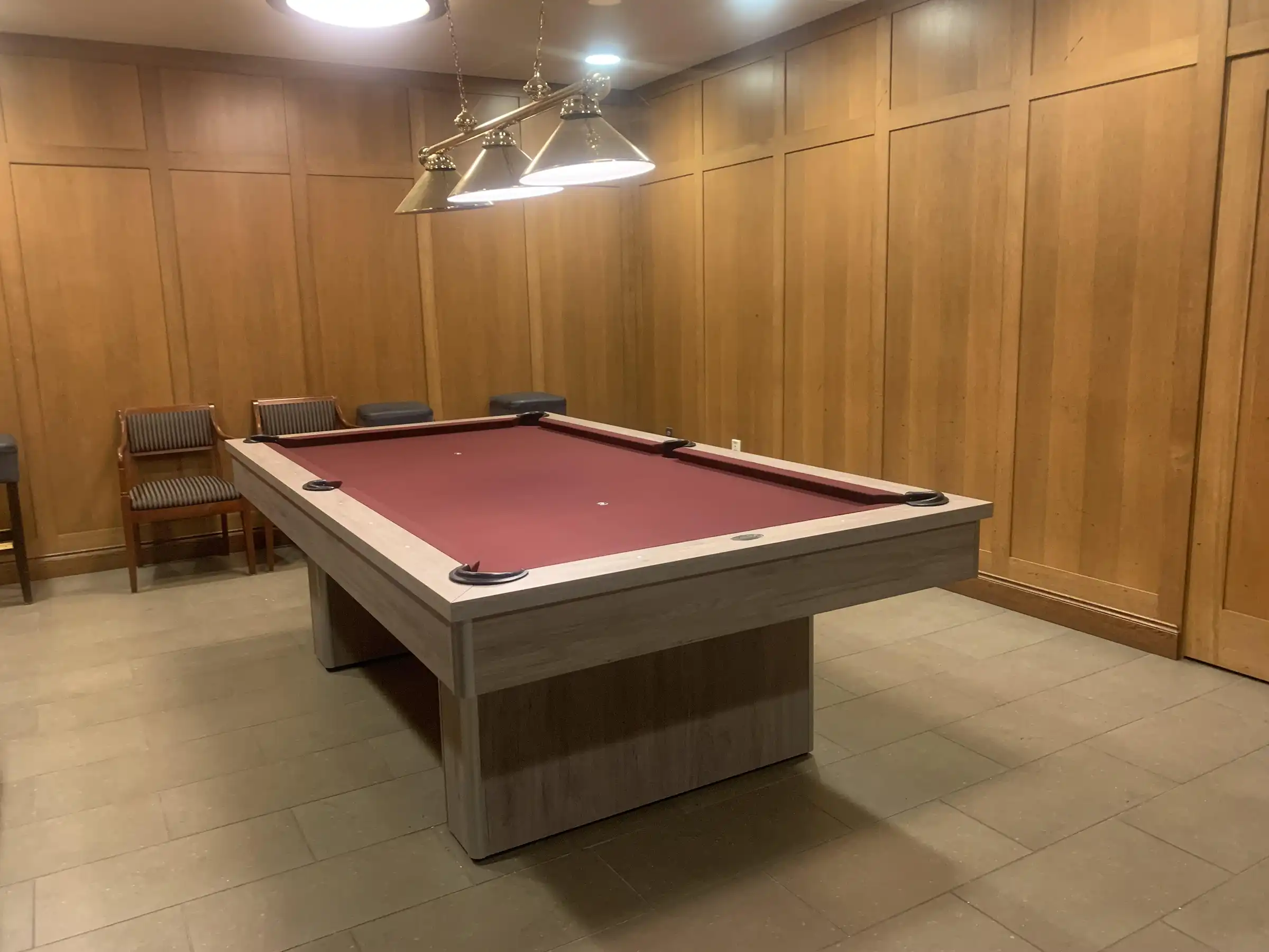 A modern pool table with maroon red felt and gray wooden base sits in a wood-paneled room with overhead lighting and striped chairs along the wall.